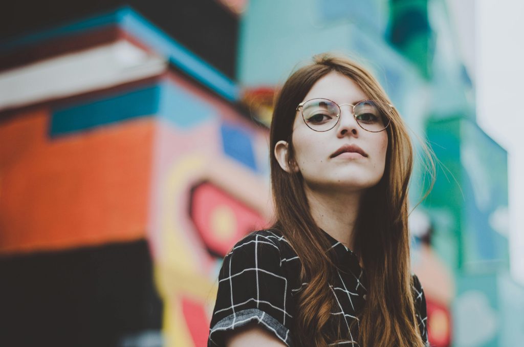 Portrait of a fashionable woman with glasses posing confidently outdoors in front of a vibrant mural.